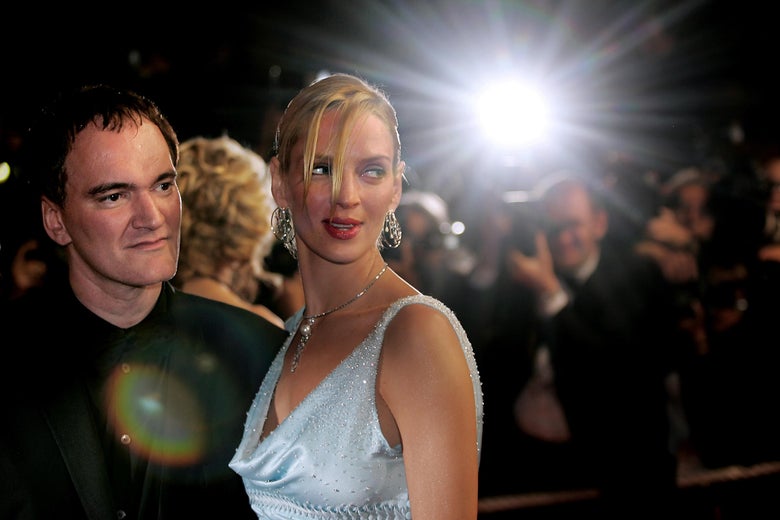 Quentin Tarantino and Uma Thurman stand on a red carpet. In the background, a photographer's bulb flashes.